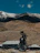 A rider on a motorcycle pauses on a mountain overlook with expansive Colorado peaks in the background under a clear blue sky.