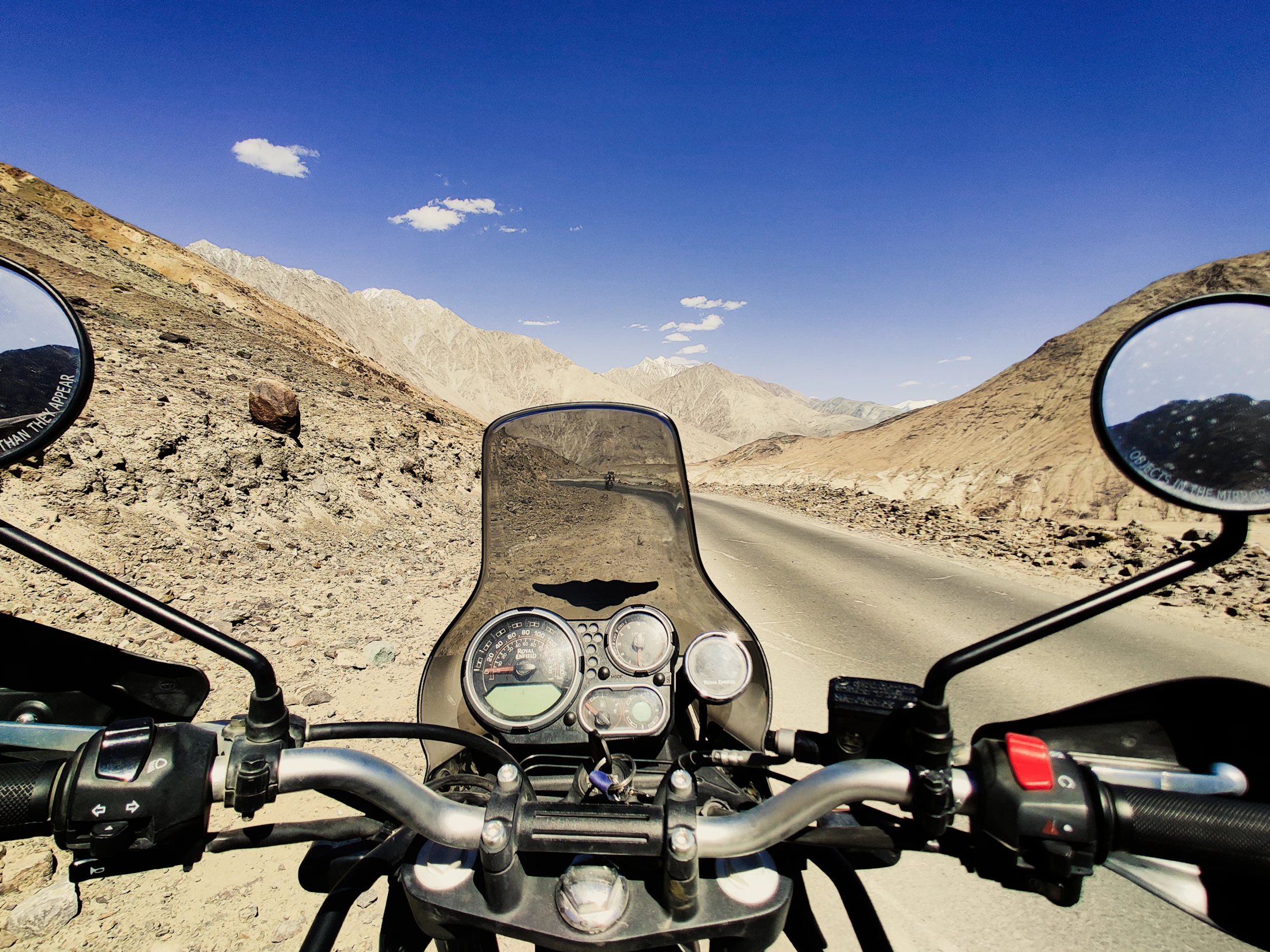 A rider's view over motorcycle handlebars down an empty high-desert road in Ladakh