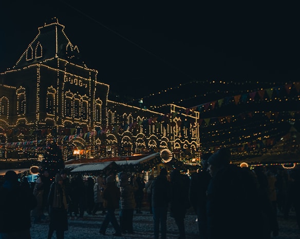 A nighttime scene at a festive market with a large, ornate building adorned with glowing string lights. A crowd of people is gathered, bundled in winter clothing, walking around under colorful triangular flags. Snow appears to cover the ground, and the atmosphere is lively and bustling with holiday decorations, including a decorated Christmas tree.