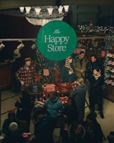 A bustling store interior during the holiday season features a central green sign with 'The Happy Store' in white text. Mannequins in plaid attire are surrounded by festive decorations and merchandise such as Christmas sweaters and accessories. Customers, bundled in winter attire, engage in shopping and browsing around a table adorned with twinkling lights and festive items.