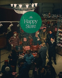 A bustling store interior during the holiday season features a central green sign with 'The Happy Store' in white text. Mannequins in plaid attire are surrounded by festive decorations and merchandise such as Christmas sweaters and accessories. Customers, bundled in winter attire, engage in shopping and browsing around a table adorned with twinkling lights and festive items.