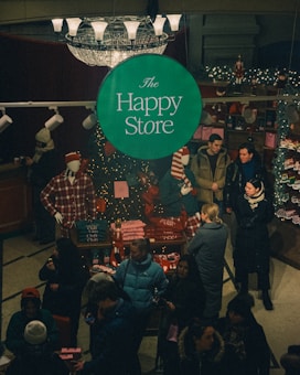 A bustling store interior during the holiday season features a central green sign with 'The Happy Store' in white text. Mannequins in plaid attire are surrounded by festive decorations and merchandise such as Christmas sweaters and accessories. Customers, bundled in winter attire, engage in shopping and browsing around a table adorned with twinkling lights and festive items.