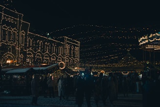 A festive night scene with brightly lit buildings and string lights hanging in the background. People are gathered, possibly at a holiday market or carnival, dressed in winter clothing. The atmosphere appears lively and vibrant with the lights creating a warm and inviting glow.
