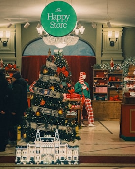 An indoor holiday display featuring a Christmas tree adorned with lights, red bows, and ornaments, positioned in a festive retail environment. A mannequin dressed in Christmas-themed attire stands nearby. Shelves in the background are filled with holiday merchandise. Overhead, a sign reads 'The Happy Store.' The lighting suggests a warm and inviting atmosphere.