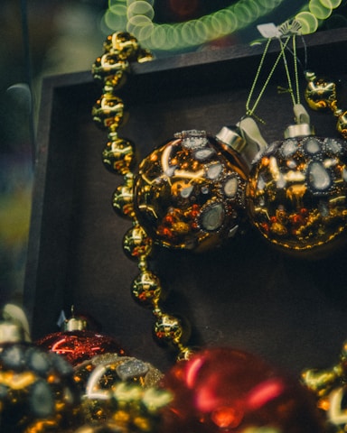 A close-up of decorative Christmas ornaments, including shiny golden and red baubles adorned with intricate patterns and glitter. The ornaments are intertwined with a beaded garland, creating a festive and luxurious atmosphere with a blurred, bokeh background.