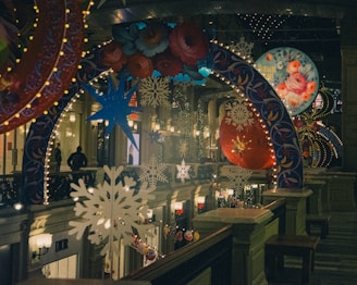 A festive indoor scene with colorful, illuminated decorations. The space is adorned with large hanging ornaments, snowflakes, and floral patterns, all lit by string lights. People can be seen in the distance, adding to the bustling atmosphere.