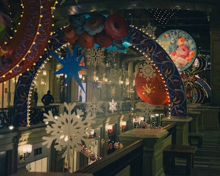 A festive indoor scene with colorful, illuminated decorations. The space is adorned with large hanging ornaments, snowflakes, and floral patterns, all lit by string lights. People can be seen in the distance, adding to the bustling atmosphere.