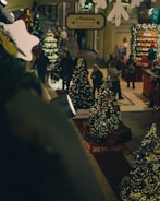 A cozy indoor shopping area decorated for the Christmas season, featuring multiple decorated Christmas trees with lights. Several people are walking around, suggesting a busy and festive atmosphere. The space includes Christmas displays, lighting decorations, and a counter with holiday-themed items.