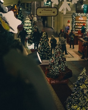 A cozy indoor shopping area decorated for the Christmas season, featuring multiple decorated Christmas trees with lights. Several people are walking around, suggesting a busy and festive atmosphere. The space includes Christmas displays, lighting decorations, and a counter with holiday-themed items.