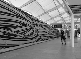 A black and white photograph of a modern station interior with an artistic wall mural featuring swirling lines. A man wearing a backpack is walking through the space, which is covered by a large, geometric ceiling with natural light streaming through.