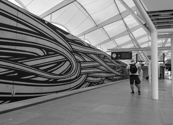 A black and white photograph of a modern station interior with an artistic wall mural featuring swirling lines. A man wearing a backpack is walking through the space, which is covered by a large, geometric ceiling with natural light streaming through.