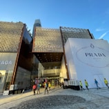 A modern building with a prominent facade featuring a large sign for Prada Milano on the right side and smaller signs for Balenciaga on the left. The ground area in front of the building is paved, and there are several workers in reflective vests and helmets walking across. Clear blue sky with a tall skyscraper in the background is visible.