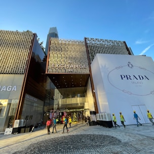 A modern building with a prominent facade featuring a large sign for Prada Milano on the right side and smaller signs for Balenciaga on the left. The ground area in front of the building is paved, and there are several workers in reflective vests and helmets walking across. Clear blue sky with a tall skyscraper in the background is visible.