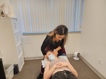A woman with glasses is performing a treatment or examination on a person lying down on a table in an office setting. The person on the table is wearing a mask and appears to be receiving some form of therapy. The room is neutral in color with vertical blinds covering a window, and there is a small fan on the floor.