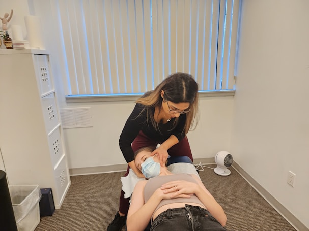 A woman with glasses is performing a treatment or examination on a person lying down on a table in an office setting. The person on the table is wearing a mask and appears to be receiving some form of therapy. The room is neutral in color with vertical blinds covering a window, and there is a small fan on the floor.