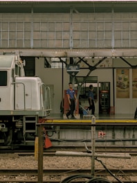 A train station platform with a partially visible train on the left. Two people in uniform are standing on the platform, which features a sign indicating 'Jalur Track 4'. The background shows the station with advertisements and the roof supported by metal beams.