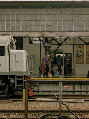 A train station platform with a partially visible train on the left. Two people in uniform are standing on the platform, which features a sign indicating 'Jalur Track 4'. The background shows the station with advertisements and the roof supported by metal beams.