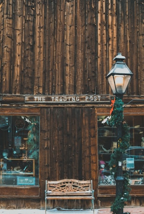 A wooden storefront with a rustic appearance features two display windows and a sign reading 'The Trading Post.' A decorative bench with 'Welcome' carved into it sits beneath the sign. A vintage-style street lamp adorned with holiday garland stands nearby, adding a festive touch. The ground is lightly dusted with snow, providing a cozy, wintry atmosphere.