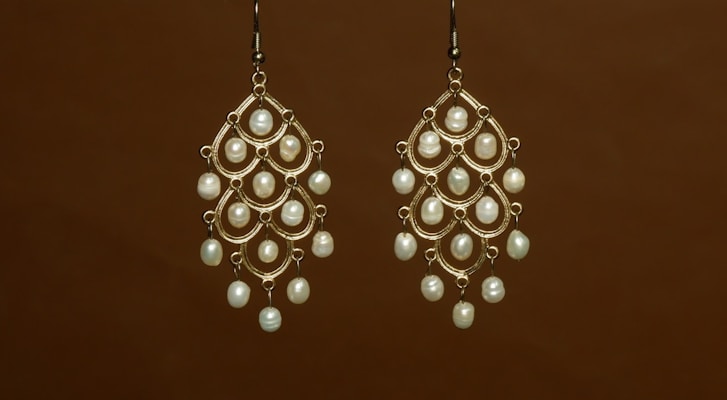 Two intricately designed chandelier earrings feature multiple white pearls set in a gold frame against a plain brown background.