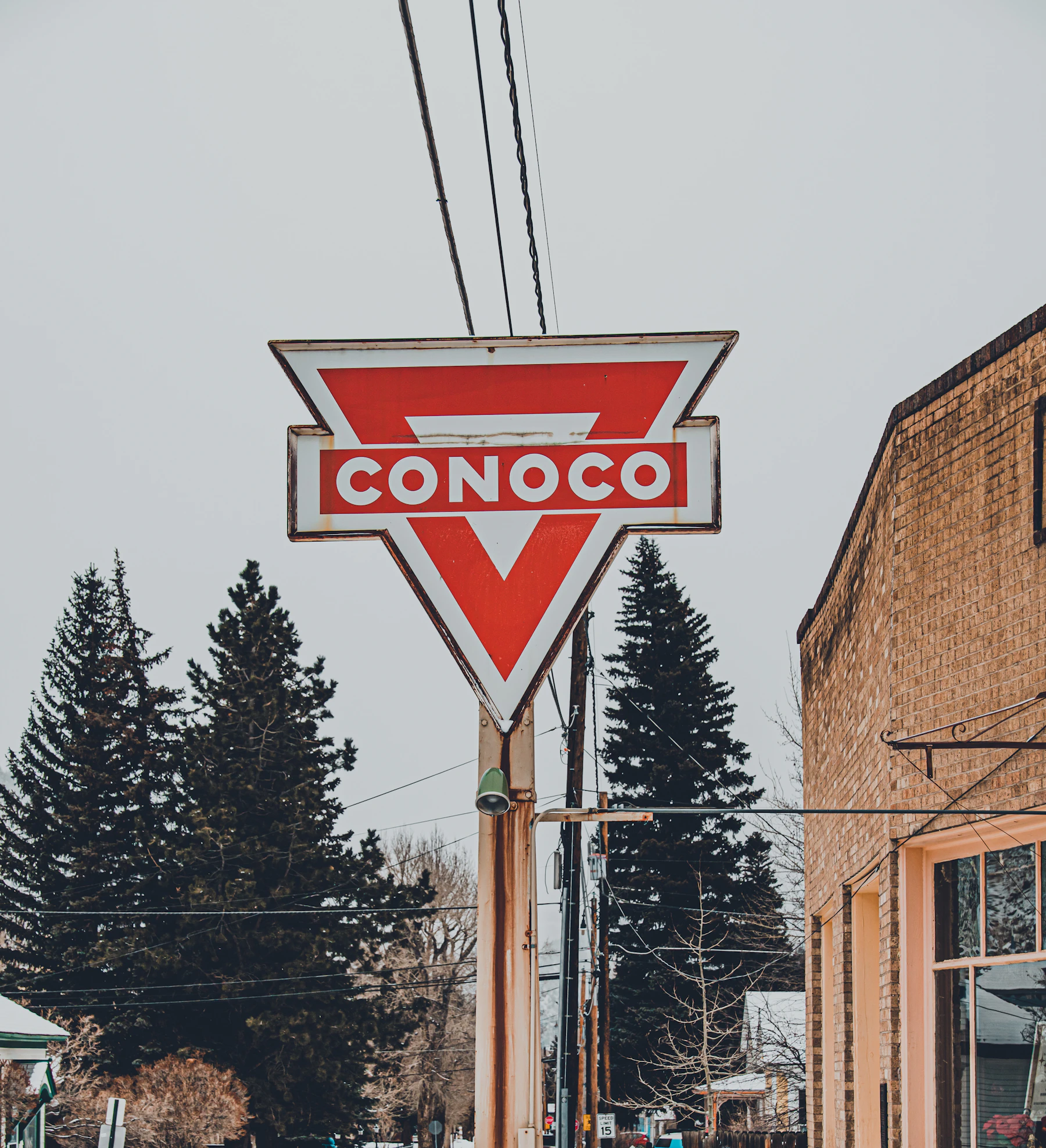 A red and white triangular Conoco sign on a pole beside a brick building, pine trees behind