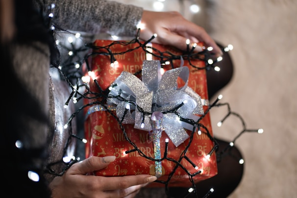 A person holding a festive wrapped gift adorned with shiny silver ribbon and surrounded by glowing string lights. The gift wrapping is predominantly red with holiday-themed text and illustrations, including ornaments and phrases of joy. The scene suggests warmth and holiday celebration.