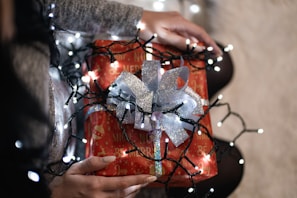 A person holding a festive wrapped gift adorned with shiny silver ribbon and surrounded by glowing string lights. The gift wrapping is predominantly red with holiday-themed text and illustrations, including ornaments and phrases of joy. The scene suggests warmth and holiday celebration.