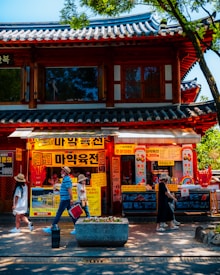 A traditional Korean building with colorful signage and advertisements. Several people are walking on the sidewalk, some wearing sun hats. The building has a tiled roof and wooden architectural elements, and the area is shaded by trees.