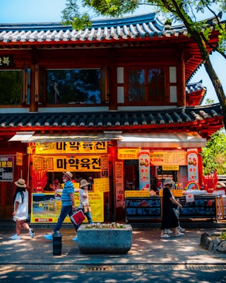 A traditional Korean building with colorful signage and advertisements. Several people are walking on the sidewalk, some wearing sun hats. The building has a tiled roof and wooden architectural elements, and the area is shaded by trees.