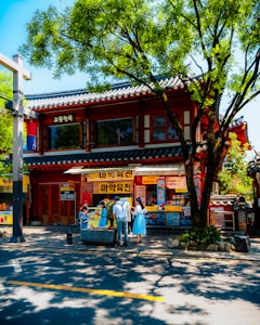 A traditional Korean building with intricate wooden designs and a tiled roof is situated among lush green trees. In front of the building, several people are gathered near a planter, some of whom are wearing blue hanbok, a traditional Korean garment. The building is adorned with bright yellow signs featuring Korean text. The scene is lively and colorful, with sunlight creating dappled shadows on the street below.