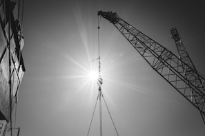 A black and white image depicting a construction scene with a crane lifting materials against the bright sun. The sunlight creates a starburst effect, emphasizing the geometric patterns of the crane's structure. On the left, part of a building is visible in shadow.