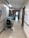 A hospital hallway with white walls, fluorescent lighting, and a polished floor. There are medical carts and equipment lining the walls, along with handrails and informational signage. The corridor is empty, leading towards a doorway with visible signage above the door.