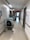 A hospital hallway with white walls, fluorescent lighting, and a polished floor. There are medical carts and equipment lining the walls, along with handrails and informational signage. The corridor is empty, leading towards a doorway with visible signage above the door.