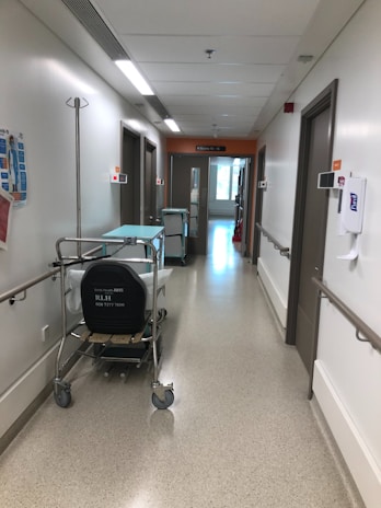 A hospital hallway with white walls, fluorescent lighting, and a polished floor. There are medical carts and equipment lining the walls, along with handrails and informational signage. The corridor is empty, leading towards a doorway with visible signage above the door.