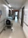 A hospital hallway with white walls, fluorescent lighting, and a polished floor. There are medical carts and equipment lining the walls, along with handrails and informational signage. The corridor is empty, leading towards a doorway with visible signage above the door.