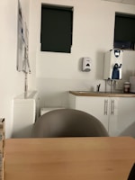 The image depicts a small office or break room setting featuring a wooden table with a chair, a countertop with a sink, a wall-mounted soap dispenser, and two windows with dark green blinds. Some papers are attached to the wall, and a water dispenser is present on the counter.