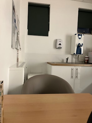 The image depicts a small office or break room setting featuring a wooden table with a chair, a countertop with a sink, a wall-mounted soap dispenser, and two windows with dark green blinds. Some papers are attached to the wall, and a water dispenser is present on the counter.