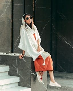 A person wearing a long white shawl over an orange dress is sitting on a marble ledge. They have long dark hair, sunglasses, and are smiling. The background features a dark marble texture with white veining.