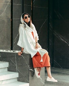 A person wearing a long white shawl over an orange dress is sitting on a marble ledge. They have long dark hair, sunglasses, and are smiling. The background features a dark marble texture with white veining.
