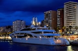 A luxurious white yacht is docked in a bustling marina, with towering modern apartment buildings and palm trees in the background. The night sky is dark blue, and the lights from the buildings and the yacht create a vibrant scene. Some buildings have balconies, and there is a sense of activity and nightlife around the marina area.