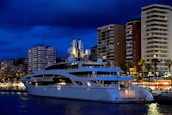 A luxurious white yacht is docked in a bustling marina, with towering modern apartment buildings and palm trees in the background. The night sky is dark blue, and the lights from the buildings and the yacht create a vibrant scene. Some buildings have balconies, and there is a sense of activity and nightlife around the marina area.