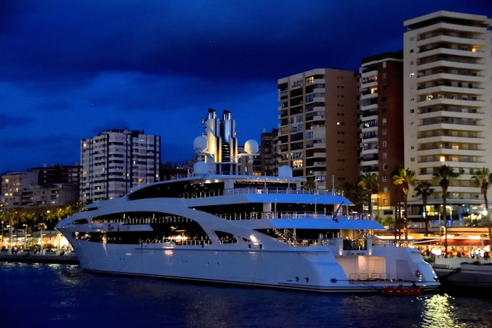 A luxurious white yacht is docked in a bustling marina, with towering modern apartment buildings and palm trees in the background. The night sky is dark blue, and the lights from the buildings and the yacht create a vibrant scene. Some buildings have balconies, and there is a sense of activity and nightlife around the marina area.