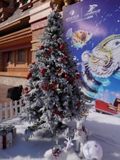 A snow-covered Christmas tree decorated with red and gold ornaments stands in front of a themed backdrop. Beside the tree, there are two small white reindeer decorations and a wrapped gift on artificial snow.