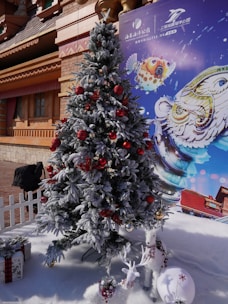 A snow-covered Christmas tree decorated with red and gold ornaments stands in front of a themed backdrop. Beside the tree, there are two small white reindeer decorations and a wrapped gift on artificial snow.