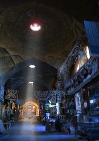 A dimly lit bazaar corridor filled with traditional textiles, lanterns, and various goods hanging from the walls and ceilings. The arched architecture creates an intimate and historic atmosphere. Sunlight streams in, casting a blue hue over the surroundings, while the high dome ceiling enhances the feeling of depth.