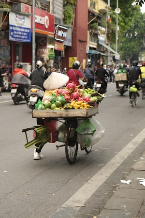 A bustling street scene features a bicycle vendor transporting a variety of fresh produce including fruits and vegetables. The vendor wears a traditional conical hat and is surrounded by motorcyclists. Street signs and shops are visible in the background.