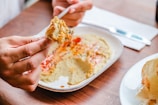 A person is holding pita bread with a portion of hummus on it, above a plate of hummus garnished with paprika and olive oil. The setting appears casual with a wooden table, and there is a knife and a napkin in the background.