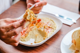 A person is holding pita bread with a portion of hummus on it, above a plate of hummus garnished with paprika and olive oil. The setting appears casual with a wooden table, and there is a knife and a napkin in the background.