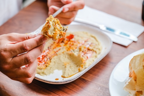 A person is holding pita bread with a portion of hummus on it, above a plate of hummus garnished with paprika and olive oil. The setting appears casual with a wooden table, and there is a knife and a napkin in the background.