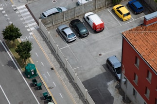 An aerial view of a parking lot with several cars parked in designated spots. The lot is adjacent to a building with a terracotta roof. A street runs alongside the parking lot, featuring bicycle lanes and waste bins lined up near the sidewalk. Trees border the road, providing green accents to the scene.