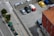 An aerial view of a parking lot with several cars parked in designated spots. The lot is adjacent to a building with a terracotta roof. A street runs alongside the parking lot, featuring bicycle lanes and waste bins lined up near the sidewalk. Trees border the road, providing green accents to the scene.